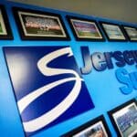 JerseySurf A blue wall displays a large Jersey Surf logo and is decorated with framed group photos—possibly including Jersey Surf new board members—arranged in rows, showcasing different years of a marching group or team.