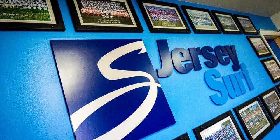 JerseySurf A blue wall displays a large Jersey Surf logo and is decorated with framed group photos—possibly including Jersey Surf new board members—arranged in rows, showcasing different years of a marching group or team.