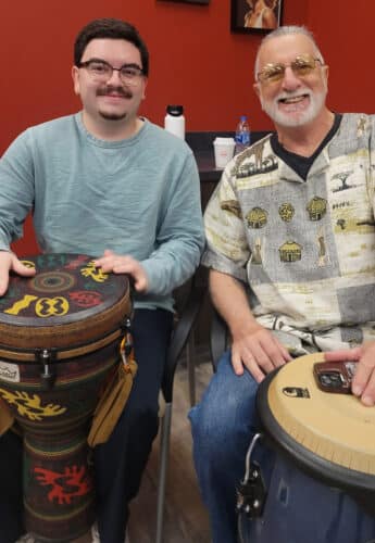 JerseySurf Two men sit side by side indoors, both smiling at the camera. At this Mobile Music Lab session, one holds a colorful djembe drum and the other a conga drum, creating a vibrant scene of Village Music Circles and shared rhythm.