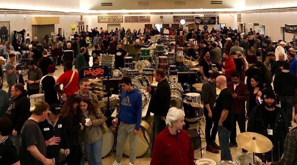 JerseySurf A large crowd gathers indoors at the Jersey Surf Delaware Drum Show, surrounded by numerous drum sets and cymbals on display. Attendees are talking, browsing, and inspecting the musical instruments.