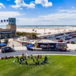JerseySurf Aerial view of a music event near a beach, with people gathered on grass, colorful tents, a MUSIC LAB truck, parking lot full of cars, and a modern building labeled CONVENTION CENTER in the background.