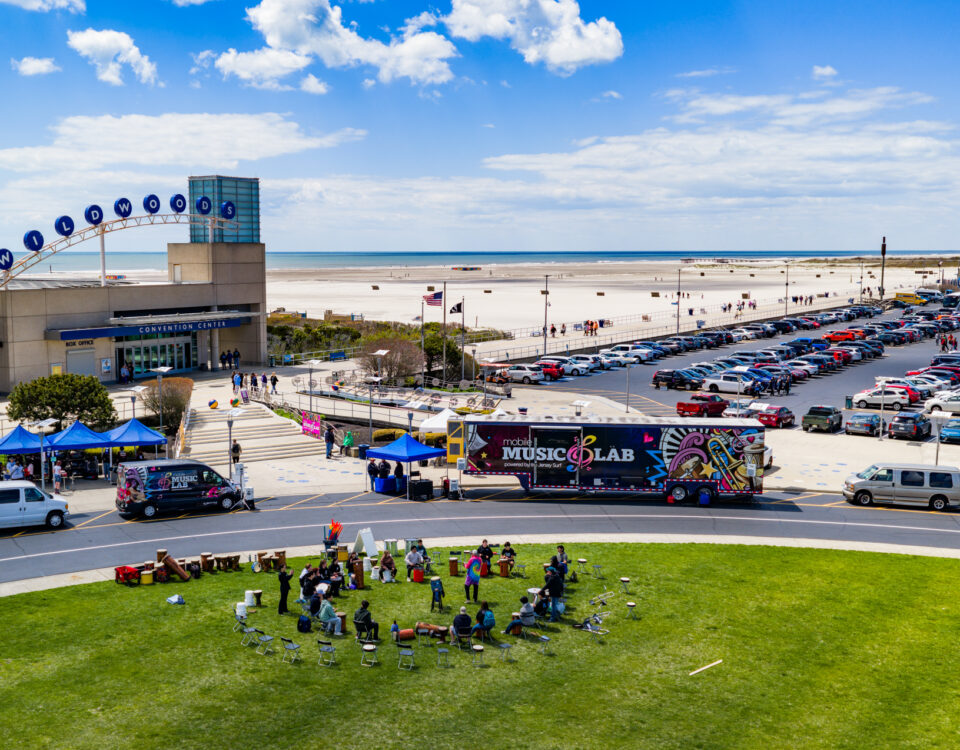 JerseySurf Aerial view of a music event near a beach, with people gathered on grass, colorful tents, a MUSIC LAB truck, parking lot full of cars, and a modern building labeled CONVENTION CENTER in the background.