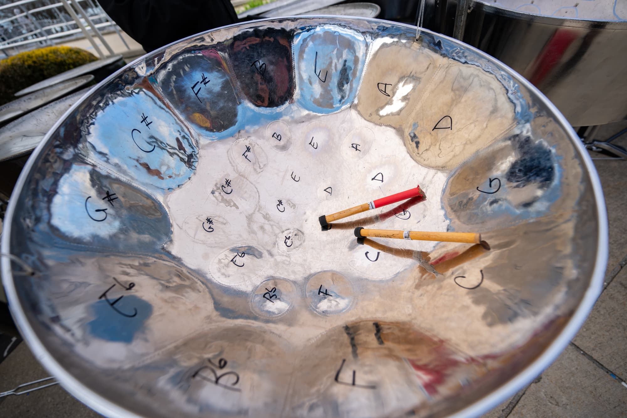 JerseySurf Close-up of a steelpan drum with musical notes marked on its surface. Two wooden mallets with colored handles rest inside the drum, reflecting the outdoor light.